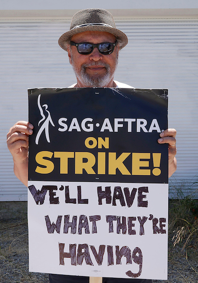 Actor Billy Crystal in a hat and sunglasses holding a sign that says "SAG AFTRA on Strike! We'll have what they're having"