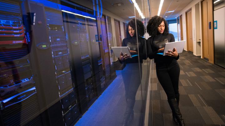 A woman on her laptop leaning against a glass wall next to a server room.