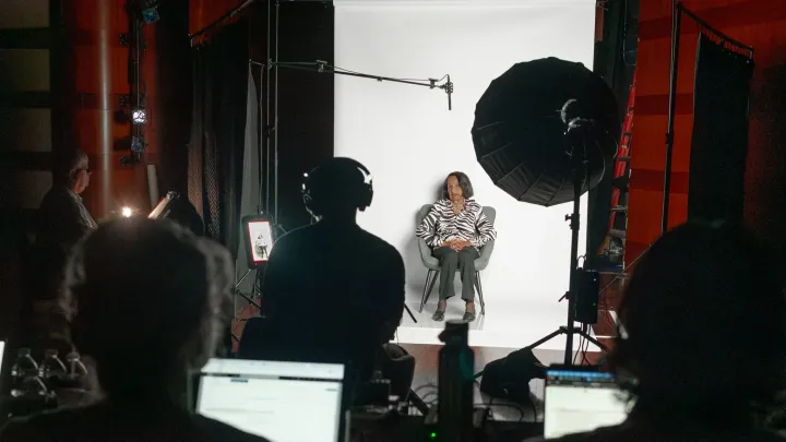 Holocaust survivor Celia Kenner sits for her Interactive Interview at the Museum of Jewish Heritage in front of a white backdrop with a light, microphone, camera, and table of staff at laptops in front of her.