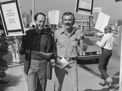 A black & white photo of William Schallert on the left holding a picket sign that says "SAG Protest against AMPTP. Women and Minorities in television: Window dressing on the set" and Dennis Weaver on the right holding a sign that says "SAG protest against AMPTP. Women & minorities: Not seen on the American scene" as they are on the picket line in front of the Academy of Motion Picture and Television producers' offices in Los Angeles, Calif.