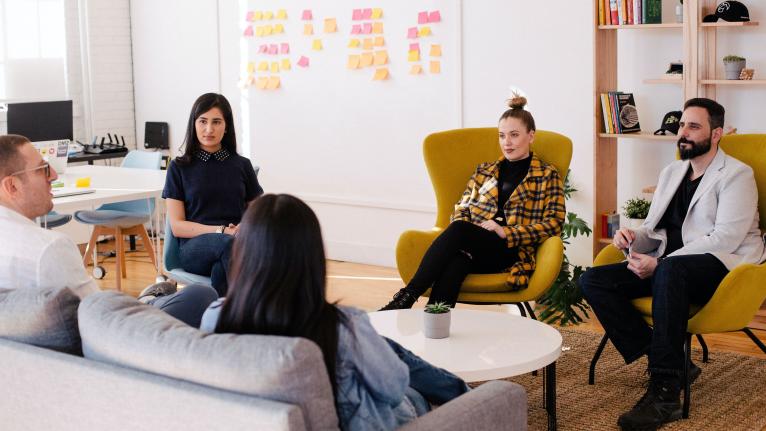 A group of people meeting in a small office sitting on a couch and chairs.